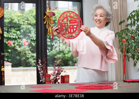Sorridente donna cinese anziana in piedi a tavola con carta rossa per il Capodanno cinese tagliata a casa Foto Stock