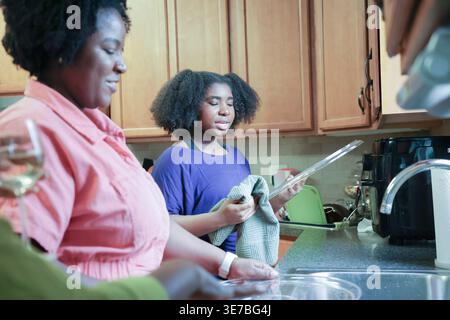 La figlia nera con afro che usa un asciugamano da cucina nelle piastre di asciugatura aiutando sua madre mentre si trova vicino al lavandino della cucina a casa Foto Stock
