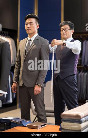 Stilisti cinesi senior che misurano i clienti cinesi maschi mentre si trovano in una boutique di moda Foto Stock