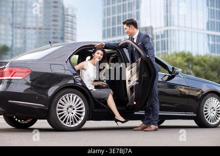 Donna d'affari cinese che scende dall'auto con autista maschio in piedi e tiene la porta sulla strada della città al tramonto Foto Stock