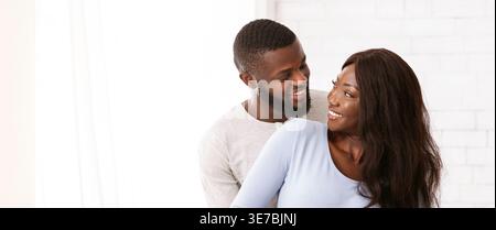 Uomo nero sorridente che abbraccia moglie incinta da dietro guardandosi l'un l'altro mentre si trova in camera da letto al chiuso Foto Stock