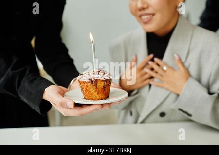 Donna asiatica che dà cupcake con candela alla collega donna che celebra il compleanno seduto al tavolo in ufficio Foto Stock