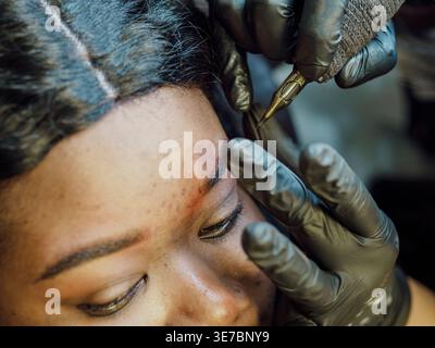 Mani di cosmetologa ispanica donna che indossa guanti applicazione permanente trucco tatuaggio sulle sopracciglia di cliente donna nera che utilizza macchina tatuaggio in s... Foto Stock