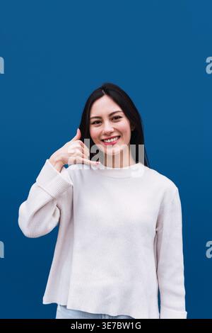 Donna asiatica sorridente che mi fa un gesto della mano in piedi su sfondo blu Foto Stock