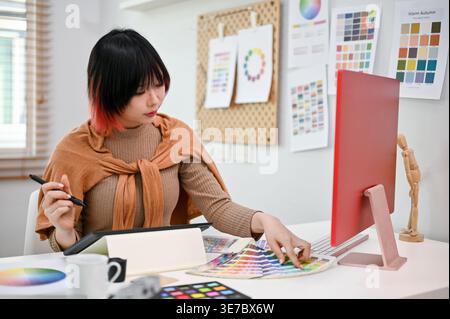 Donna asiatica che tiene la penna stilo al lavoro sul suo progetto mentre si siede a tavola in ufficio Foto Stock