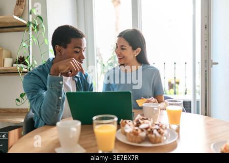 Sorridente coppia ispanica e caucasica che fa shopping online con un portatile mentre si siede a tavola con pasticcini e succo di frutta a casa Foto Stock