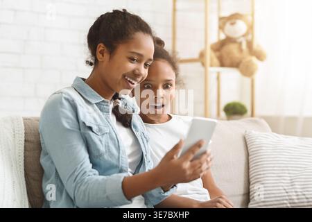 Adolescente nera che mostra lo smartphone alla sorella minore seduta sul divano in salotto di casa Foto Stock