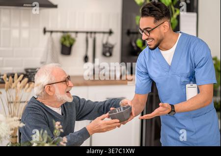 Sorridente assistente maschio arabo in occhiali da vista che dà cibo in piedi vicino a un uomo anziano seduto a tavola a casa Foto Stock