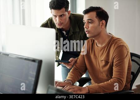 Programmatori maschi indiani che tengono il tablet appoggiato in avanti accanto al collega maschio indiano che digita i codici sul computer in ufficio Foto Stock