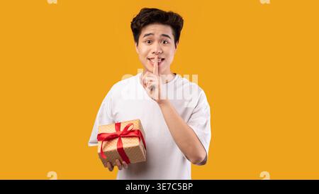 Uomo asiatico che indossa una camicia bianca facendo segno di silenzio e tenendo in mano una scatola regalo con nastro rosso in piedi contro la parete arancione in studio Foto Stock