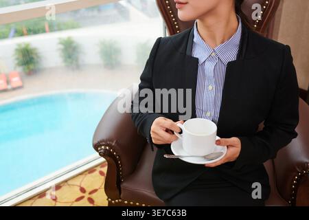 Donna d'affari cinese che guarda attraverso la finestra tenendo una tazza di caffè seduto sulla sedia nella hall dell'hotel Foto Stock