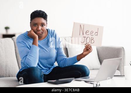 Donna nera con afro corto che tiene in mano un cartello di cartone con il testo del lavoro da lavorare mentre si siede sul divano con la testa appoggiata a mano nel soggiorno di casa Foto Stock