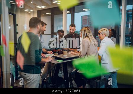 Uomo d'affari di colore che tiene in mano la festa del telefono con i colleghi intorno al tavolo in ufficio Foto Stock