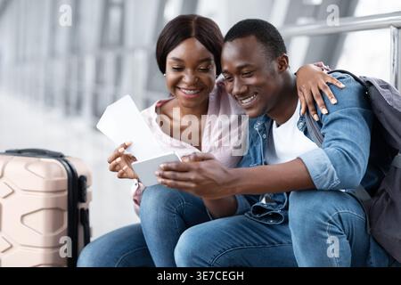 Sorridente viaggiatore di coppia nera con biglietti aerei tramite smartphone mentre si siede sul passaggio pedonale dell'aeroporto Foto Stock