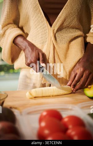 Le mani di Black Woman affettano la banana in pezzi con un coltello sul tagliere in piedi al bancone della cucina di casa Foto Stock