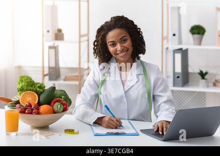 Nutrizionista di colore che lavora al computer portatile e scrive sugli appunti mentre si siede al tavolo con una ciotola di frutta e verdura in clinica Foto Stock