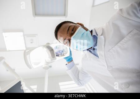 Dentista maschio nero che indossa la maschera e i guanti che accendono la lampada prima del trattamento dentale in clinica Foto Stock