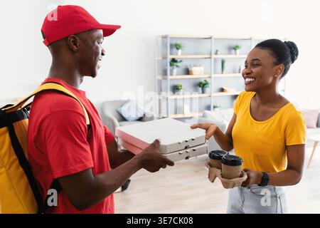 Donna nera sorridente che riceve scatole per pizza e tazze da caffè usa e getta dal fattorino mentre si trova davanti alla porta di casa Foto Stock
