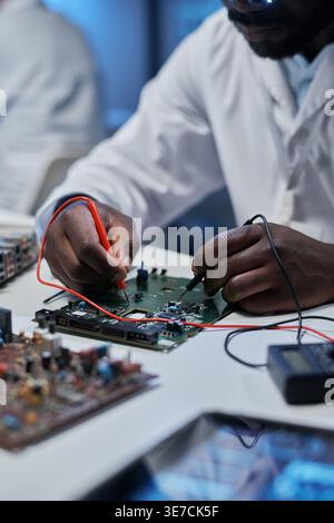 Immagine ritagliata di un ingegnere informatico maschio nero che lavora con le mani sulla scheda madre mentre si siede al tavolo in laboratorio Foto Stock