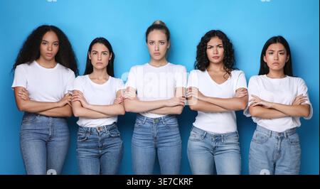 Gruppo di donne molto eterogeneo che indossano camicie e jeans in piedi con le braccia incrociate su sfondo blu in studio Foto Stock