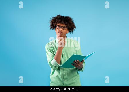 Attento studente di sesso maschile nero con capelli ricci e occhiali da vista, mento appoggiato a mano tenendo il libro davanti allo sfondo blu a st... Foto Stock