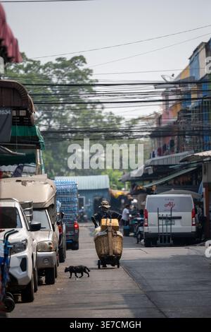 Bangkok Thailandia 16 gennaio 2026 Un lavoratore spinge un carretto carico di grandi cesti lungo una strada al mercato Khlong Toei mentre un gatto nero attraversa t Foto Stock
