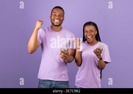 Cheerful Black coppia che tiene gli smartphone in piedi con pugni serrati su sfondo viola in studio Foto Stock