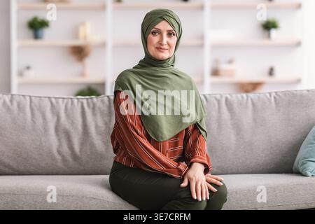 Donna musulmana mediorientale di mezza età in hijab seduta sul divano nel soggiorno di casa Foto Stock