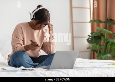 Donna nera che indossa le cuffie che toccano la testa soffre di mal di testa tenendo la penna seduta con le gambe incrociate sul letto vicino al laptop nella camera da letto di casa Foto Stock
