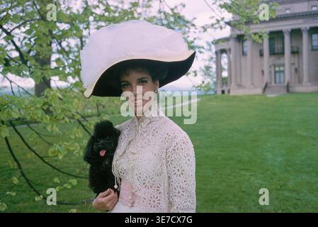 Wendy Vanderbilt posa all'aperto nella tenuta di Frederick Vanderbilt "Hyde Park", indossando un elegante abito in pizzo e un cappello a teglia larga mentre tiene in mano un piccolo cane, con la villa neoclassica visibile sullo sfondo. Fotografia di Toni Frissell per la rivista Life, 1964. Foto Stock