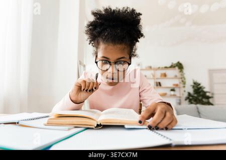Scioccata preadolescente nera che tiene un libro da lettura a penna seduto a tavola a casa Foto Stock
