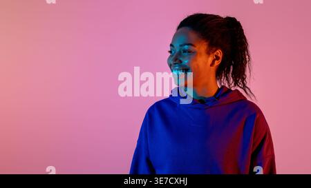Donna nera sorridente che indossa una felpa con cappuccio che guarda lontano in piedi sullo sfondo rosa in studio Foto Stock