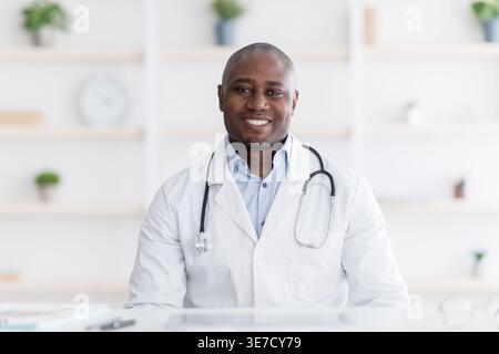 Medico nero sorridente che indossa lo stetoscopio intorno al collo seduto alla scrivania in clinica Foto Stock