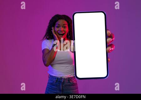 Donna nera eccitata con camicia bianca e jeans con grande smartphone con schermo bianco vuoto su sfondo viola e rosa in studio Foto Stock