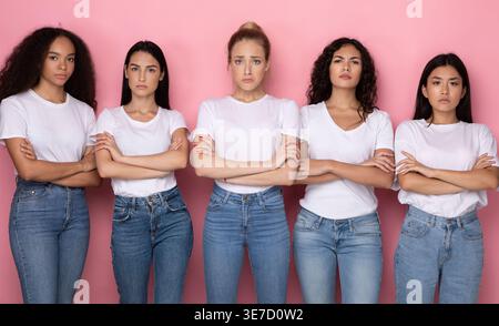Gruppo di donne molto eterogeneo che indossano camicie bianche e jeans in piedi con le braccia incrociate su sfondo rosa in studio Foto Stock