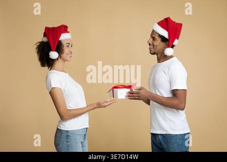 Uomo nero che indossa il cappello di Babbo Natale e regala una confezione regalo con nastro rosso alla fidanzata mentre si trova sullo sfondo marrone in studio Foto Stock