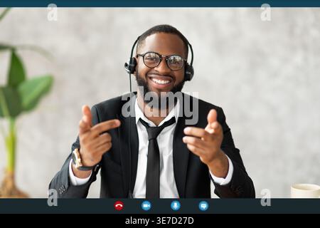 Uomo d'affari nero sorridente con le cuffie che gestiscono le mani durante le videochiamate, seduto contro una parete grigia in ufficio Foto Stock