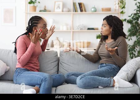 Amiche nere che discutono tra loro gestendo le mani mentre sedevano insieme sul divano nel salotto di casa Foto Stock