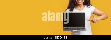 Donna nera sorridente che indossa una camicia bianca che mostra un laptop mentre si trova sullo sfondo giallo in studio Foto Stock