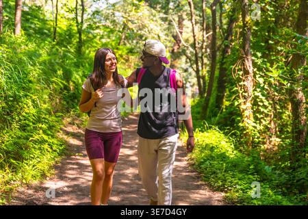 Coppia nera e caucasica con zaini che parlano tra di loro mentre camminano nella foresta nelle giornate di sole Foto Stock