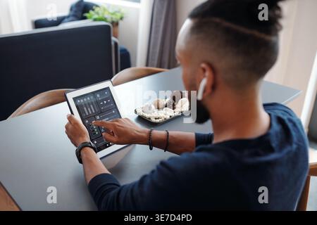Vista posteriore dell'uomo mediorientale che ascolta la musica sugli auricolari e utilizza l'applicazione volume sul tablet seduto al tavolo in salotto di casa Foto Stock