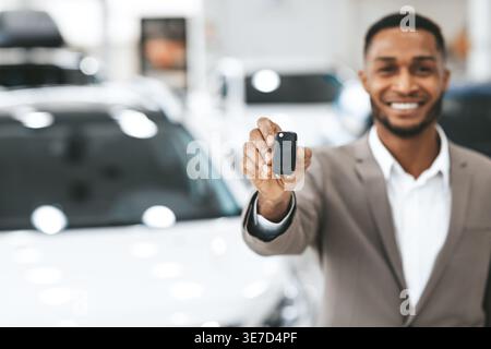 Venditore nero in tuta aziendale che mostra la chiave automatica mentre si trova davanti all'auto nello showroom Foto Stock