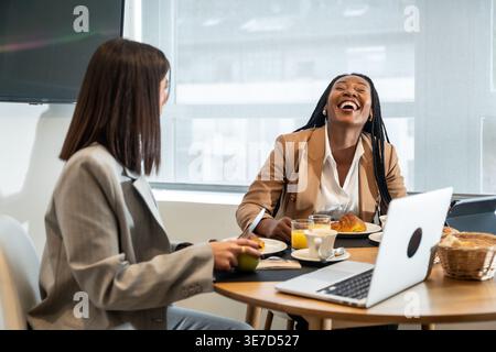 Donne d'affari nere e caucasiche che fanno colazione e parlano tra di loro mentre si siedono al tavolo con un computer portatile e un tablet in camera d'albergo Foto Stock