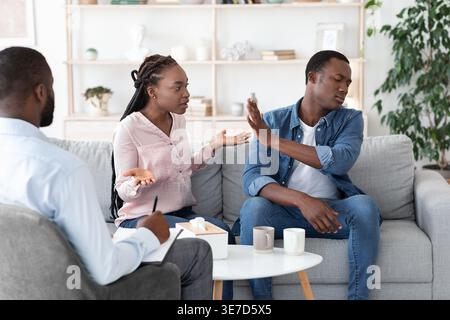 Coppia nera che litiga mentre si siede sul divano e ha una consulenza matrimoniale con un terapista di sesso maschile nero in ufficio Foto Stock