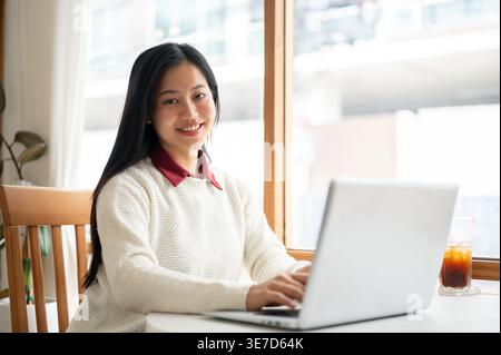 Donna tailandese sorridente seduta al tavolo con un computer portatile e un bicchiere di caffè ghiacciato nel caffè Foto Stock