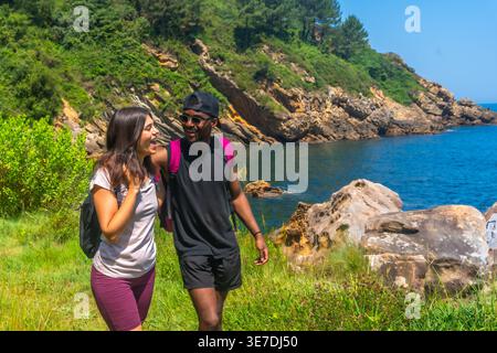 Coppia nera e caucasica con zaini che camminano lungo la costa durante il giorno di sole Foto Stock