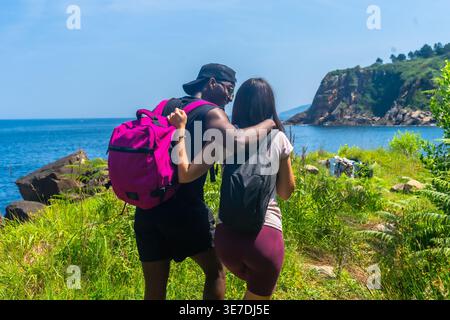 Vista posteriore della coppia nera e caucasica con zaini che camminano verso il mare nelle giornate di sole Foto Stock