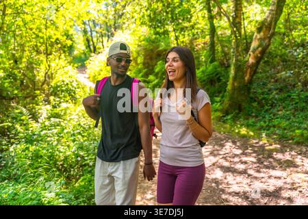 Coppia nera e caucasica con zaini che parlano tra di loro mentre camminano nella foresta nelle giornate di sole Foto Stock