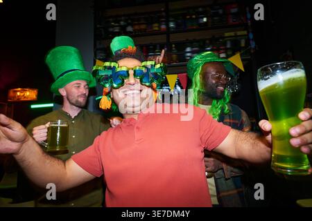 Uomo dalle corse miste con occhiali da vista in trifoglio e cappello verde che si diverte a ballare alla festa con gli amici al bar Foto Stock