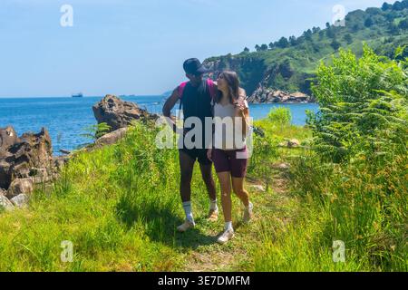 Coppia nera e caucasica con zaini che camminano lungo il sentiero costiero nelle giornate di sole Foto Stock
